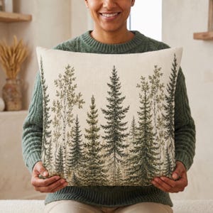 May include: A square throw pillow featuring a printed forest scene with various evergreen and deciduous trees in shades of dark green and muted brown against a light cream-colored fabric background with a visible woven texture.
