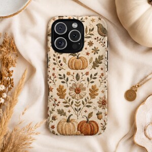 May include: A phone case featuring a cream-colored background with an embroidered-style pattern of pumpkins, flowers, leaves, acorns, and a small bird in shades of orange, brown, and green. The case has a black camera lens cutout.