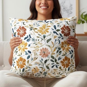 May include: A square throw pillow featuring a white background with a watercolor-style botanical print. The pattern consists of flowers and leaves in shades of mustard yellow, burnt orange, muted green, and dark blue throughout the fabric.