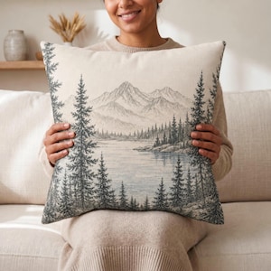 May include: A square throw pillow featuring a detailed line art illustration of a serene landscape with a calm lake, dense evergreen pine trees in the foreground, and tall mountain peaks in the background, rendered in shades of gray and cream.