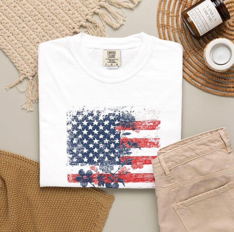 May include: White t-shirt with a distressed American flag design and floral details in red, white, and blue. The shirt is shown with beige trousers. The overall aesthetic is patriotic and casual.