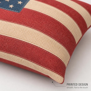 May include: A decorative pillow featuring a printed design of the American flag. The pillow has red and beige stripes, a blue corner with white stars, and the text "PRINTED DESIGN smooth, flat to the touch".