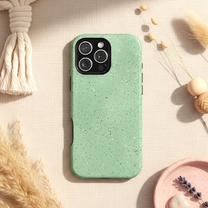 May include: A light green speckled phone case with a camera module. The case has a textured appearance and is surrounded by decorative elements, including dried plants and a pink dish.