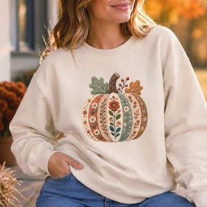 May include: A cream-colored crewneck sweatshirt featuring a central graphic of a pumpkin. The pumpkin is divided into vertical segments, each decorated with intricate floral patterns in shades of orange, green, brown, and white.