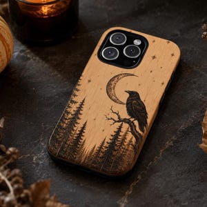 May include: A light-colored wooden phone case featuring a laser-engraved design of a raven perched on a tree branch under a crescent moon and stars, with a dense forest silhouette at the bottom, surrounding the black camera lens module.