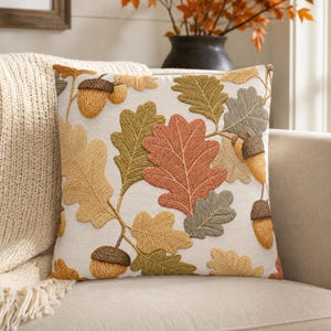 May include: A square throw pillow with an off-white base featuring an embroidered pattern of oak leaves and acorns in shades of burnt orange, olive green, mustard yellow, and brown. The design covers the front surface with detailed stitching.