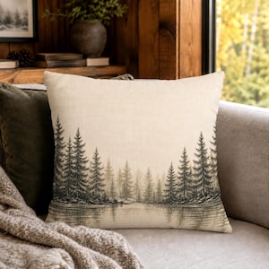 May include: A square throw pillow featuring a printed illustration of a dense pine forest bordering a calm lake. The design uses shades of dark gray and black against a light beige background, depicting trees reflected in the water.
