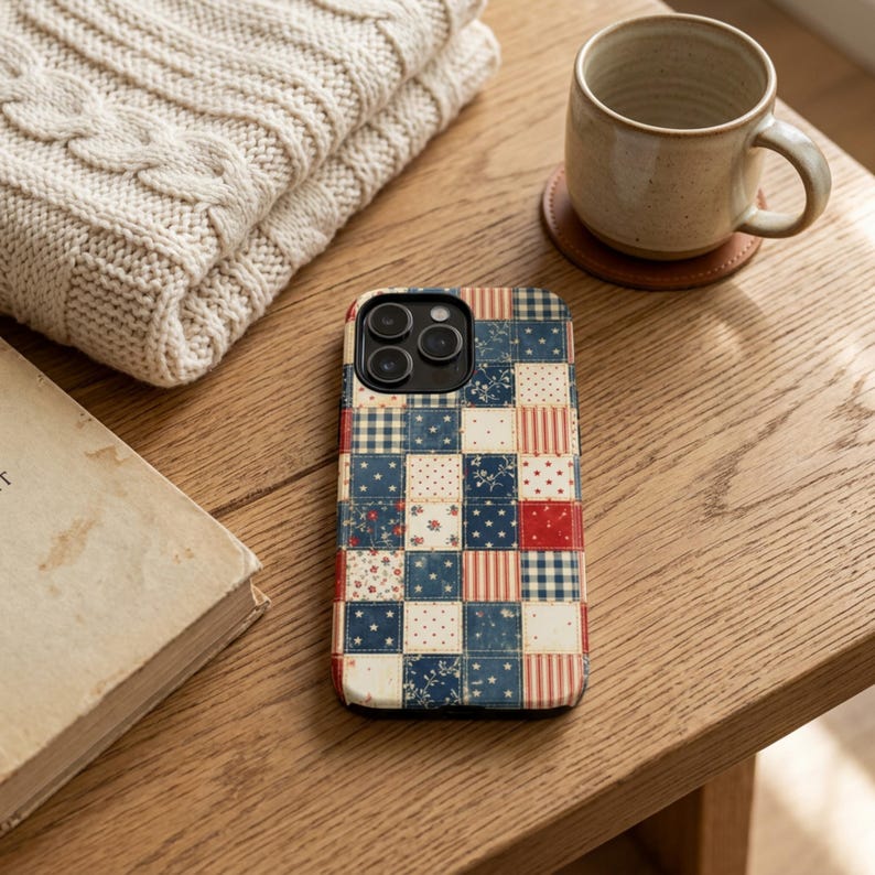 Op de afbeelding: Telefoonhoesje met een patchwork-ontwerp in rood, wit en blauw. De hoes heeft vierkanten met bloemen-, sterren- en streepmotieven. Een beige trui, een mok en een boek liggen op een houten tafel.