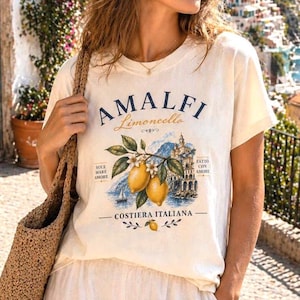 May include: Cream-colored t-shirt featuring the text "AMALFI Limoncello" above a scenic illustration of lemons, a coastal town, and the words "COSTIERA ITALIANA." The shirt has short sleeves and a classic crew neck.