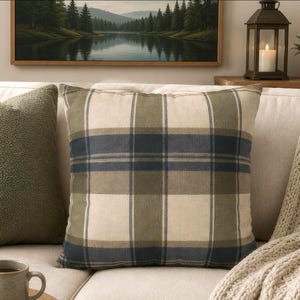 May include: A square throw pillow featuring a plaid pattern in shades of olive green, navy blue, and cream. The fabric has a visible woven texture, and the design consists of intersecting horizontal and vertical stripes forming a grid.