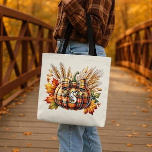 May include: A white tote bag featuring a colorful pumpkin design with a plaid bow and the letter "S". The pumpkin is surrounded by wheat and autumn leaves. The bag has black handles.