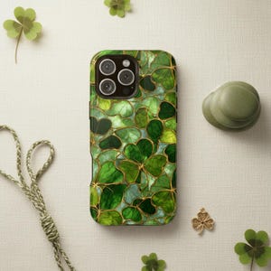 May include: A green phone case featuring a stained glass design of shamrocks with gold-colored outlines. The case has a glossy finish and a protective design. The phone case is surrounded by shamrock decorations.