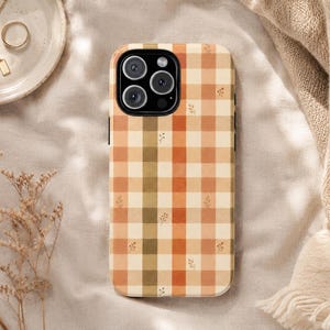May include: A rectangular smartphone case featuring a plaid pattern in shades of orange, cream, and olive green. The design includes small, delicate sprig illustrations scattered across the grid, with a black camera cutout at the top.