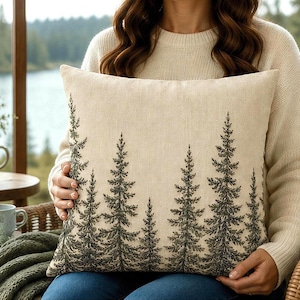 May include: A square throw pillow with a cream-colored fabric base featuring a dark green embroidered pattern of multiple pine trees. The trees vary in height and are arranged across the front surface of the pillow, creating a forest-like design.