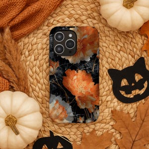 May include: A smartphone case with a floral design featuring orange and white flowers on a black background. The case has a glossy finish and a camera cutout. The case is surrounded by pumpkins, leaves, and a decorative cat.
