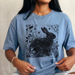 May include: A light blue t-shirt featuring a black illustration of a rabbit surrounded by flowers and foliage. The graphic is centered on the shirt. The shirt has short sleeves and a crew neck.