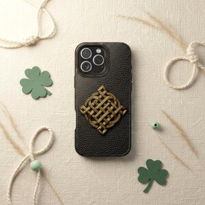 May include: A black textured phone case featuring a centered metallic gold-toned Celtic knot emblem. The case has a triple-lens camera cutout at the top. The design is surrounded by decorative green shamrock cutouts and white rope accents.