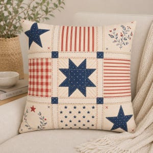 May include: A square throw pillow featuring a patchwork design with a cream background. The pattern includes navy blue stars, red and white stripes, red and white gingham checks, and small floral sprigs. The fabric has visible quilting stitches.