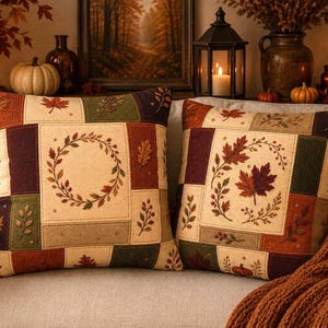 May include: Two square throw pillows featuring a patchwork design with cream, burgundy, olive green, and brown panels. The fabric displays embroidered autumn motifs including maple leaves, berry sprigs, a circular wreath, and a small pumpkin.