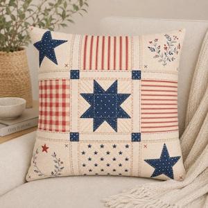 May include: Square decorative pillow with a patchwork design featuring red, white, and blue elements. The pillow has a central eight-pointed star, blue stars, red stripes, and a red and white gingham pattern. The pillow is on a white sofa.