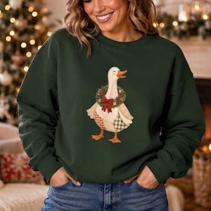 May include: A dark forest green crewneck sweatshirt featuring a central graphic of a white goose wearing a festive wreath around its neck with a red bow. The goose has a patchwork-style wing design with red and green checkered patterns.