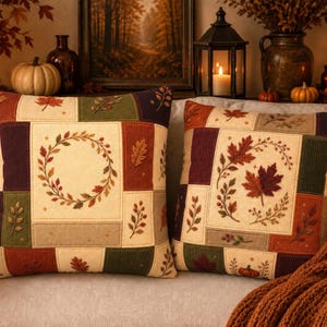 May include: Two square throw pillows featuring a patchwork design with cream, green, burgundy, and brown panels. The fabric displays embroidered autumn motifs including maple leaves, oak leaves, and circular wreaths in seasonal colors.