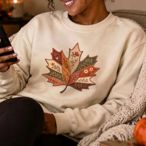 May include: A cream-colored crewneck sweatshirt featuring a large, central graphic of a maple leaf. The leaf is divided into sections, each filled with intricate, colorful floral patterns in shades of orange, red, green, and brown.