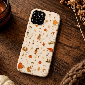 May include: A cream-colored smartphone case featuring a repeating pattern of white geese dressed in autumn-themed clothing, including sweaters, scarves, and hats, interspersed with small orange pumpkins and brown maple leaves.