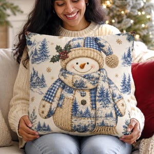 May include: A square throw pillow featuring a central snowman illustration wearing a blue and white checkered hat and scarf. The background displays a winter landscape with blue pine trees, cabins, and snowflakes on a cream fabric.