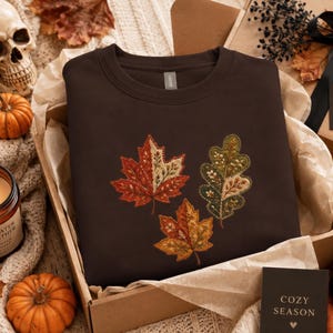 May include: A dark brown crewneck sweatshirt featuring an embroidered design of three autumn leaves in shades of red, orange, and green. The leaves display intricate stitching details. A small black card with the text COZY SEASON is included.