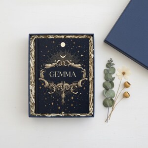 May include: A dark blue hardcover journal with the name GEMMA printed in gold, surrounded by ornate gold filigree, stars, and a crescent moon. The journal is nestled in a blue gift box filled with paper shred, accompanied by a dried eucalyptus sprig.