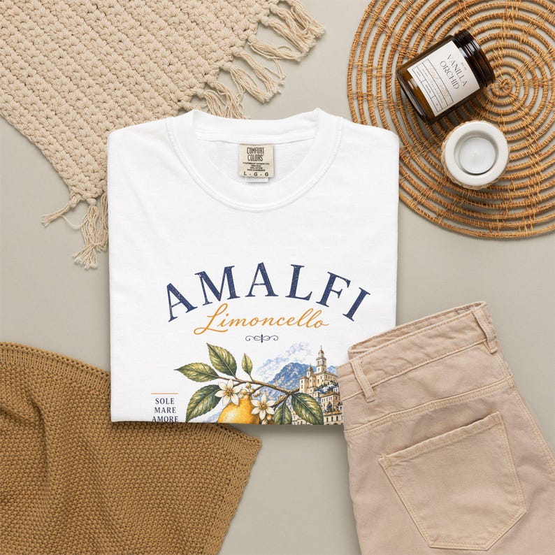 May include: White t-shirt with the word "AMALFI" in navy blue and "Limoncello" in gold script. The shirt features a lemon branch illustration with a coastal town scene. Tan pants, a woven mat, and a candle are also visible.