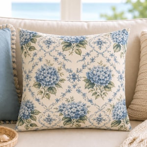 May include: A square throw pillow featuring a cream-colored background with a repeating pattern of blue hydrangea clusters, green leaves, and intricate blue scrollwork framing each floral motif in a diamond-shaped lattice arrangement.
