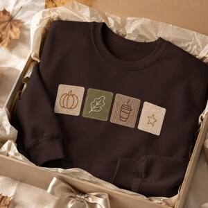 May include: A dark brown crewneck sweatshirt featuring four square embroidered patches across the chest. The patches display a pumpkin, an oak leaf, a coffee cup, and a star in shades of beige, olive green, and brown, arranged in a horizontal row.
