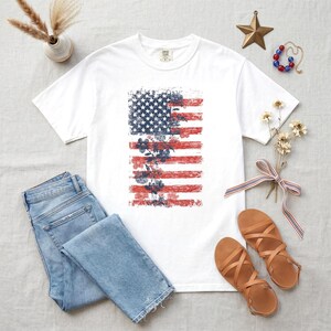 May include: White t-shirt with a distressed American flag design and floral accents. The flag design uses red and blue colours. The image also includes blue jeans and brown sandals, suggesting a casual, patriotic theme.