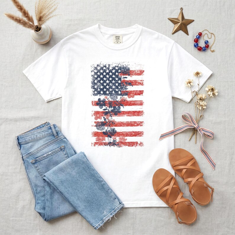 May include: White t-shirt with a distressed American flag design and floral accents. The flag design uses red and blue colours. The image also includes blue jeans and brown sandals, suggesting a casual, patriotic theme.