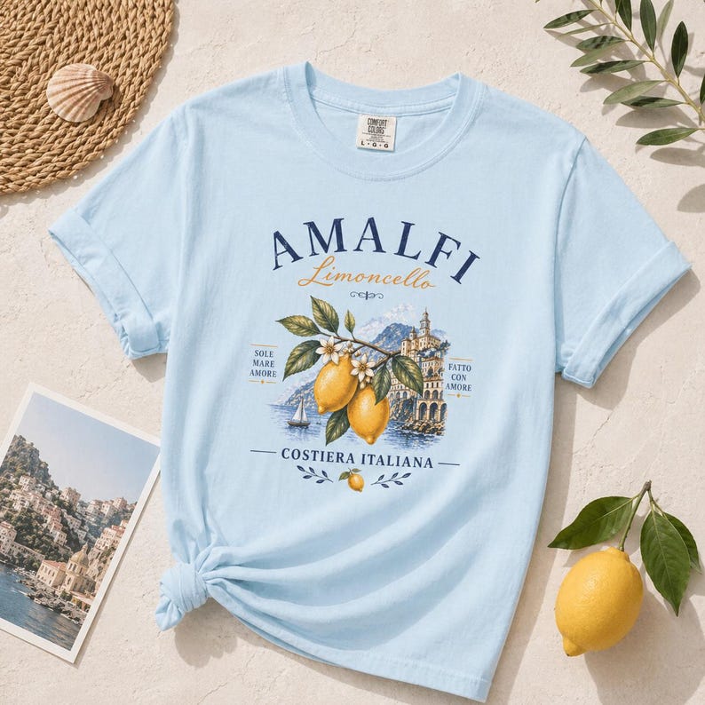 May include: Light blue t-shirt with "AMALFI Limoncello" graphic. The graphic features lemons, flowers, and a coastal Italian scene. The shirt is styled with a postcard and a lemon.