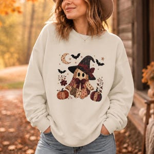 May include: A cream-colored crewneck sweatshirt featuring a central graphic of a ghost wearing a witch hat, decorated with a patchwork pattern. The design includes pumpkins, bats, a crescent moon, and floral elements in shades of brown, red, and black.