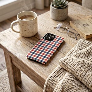 May include: A phone case with a red, white, and blue plaid pattern rests on a wooden table. A mug of coffee, glasses, a book, a succulent, and a beige knitted blanket are also on the table. The scene evokes a cozy, relaxed atmosphere.