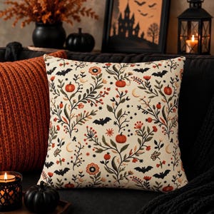 May include: A square throw pillow with a cream-colored background featuring a repeating pattern of orange pumpkins, dark green leafy vines, small red berries, black bats, and crescent moons. The design is symmetrical and covers the entire surface.