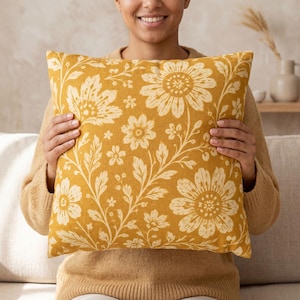 May include: A square throw pillow featuring a mustard yellow background with a repeating cream-colored floral pattern of large sunflowers, smaller blossoms, and leafy vines. The fabric has a soft, matte texture and a consistent botanical print.