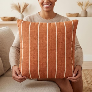 May include: A square throw pillow featuring a textured burnt orange fabric with vertical white stripes. The pillow has a woven appearance and a consistent pattern across the front surface, providing a clear view of the fabric weave and color.