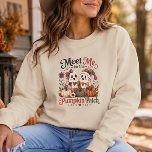 May include: A cream-colored long-sleeve sweatshirt featuring a graphic of two ghosts wearing patterned hats and bows, surrounded by pumpkins and flowers. The text reads Meet Me in the Pumpkin Patch in brown and red script and block lettering.