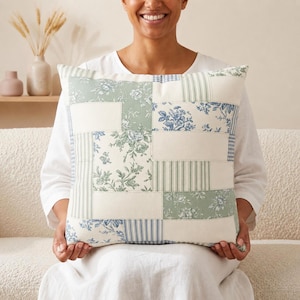May include: A square throw pillow featuring a patchwork design of cream, sage green, and blue fabric panels. The pattern includes floral toile prints, vertical stripes, and small blue and white gingham checks arranged in a rectangular grid layout.