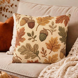 May include: A square throw pillow featuring a cream-colored background with a repeating pattern of embroidered autumn leaves in shades of orange, brown, and green, interspersed with detailed brown and green acorns throughout the design.
