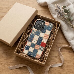 Op de afbeelding: Een telefoonhoesje met een patchwork-ontwerp in een geschenkdoos. De hoes heeft een patroon van rode, witte en blauwe vierkanten. De doos is bruin en gevuld met versnipperd papier. Een lint en gedroogde bloemen zijn ook zichtbaar.