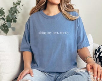 May include: A dusty blue short-sleeve crewneck t-shirt featuring the white text "doing my best. mostly." printed in a simple serif font across the center of the chest. The garment has a relaxed fit with a standard ribbed neckline.