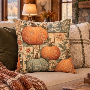May include: A square throw pillow featuring a patchwork-style print of orange and sage green pumpkins interspersed with floral motifs and leafy vines. The design includes a cream-colored background with rectangular panels containing botanical illustrations.