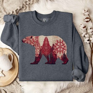 May include: A dark gray sweatshirt featuring a graphic of a bear with a red and white winter forest design. The bear's silhouette is filled with trees and snowflake patterns. The sweatshirt has long sleeves and a crew neck.