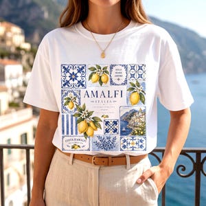 May include: White t-shirt with a colorful Amalfi, Italy design. The design features lemons, blue and white tiles, and the words "Amalfi Italia". The shirt has short sleeves and a crew neck.
